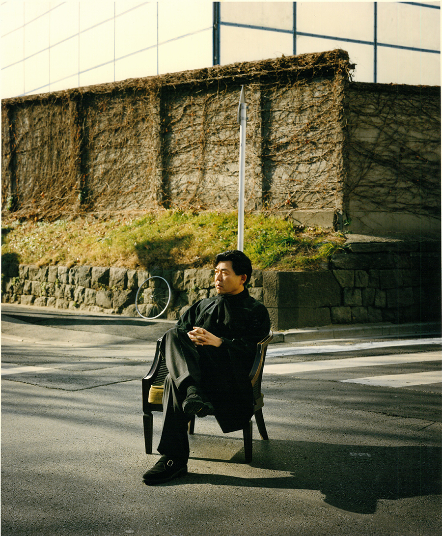 20. Arakawa sits in an armchair in the middle of an intersection in Japan. A bicycle wheel to the left and the grid lines of a building above, partially frame him in references to Modern art.